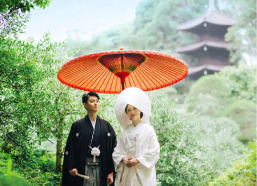 和傘を差しながら三重塔を背景に微笑む新郎新婦